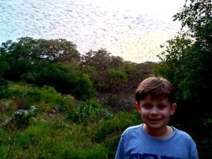 young boy in front of a cave with bats flying out of it at sunset