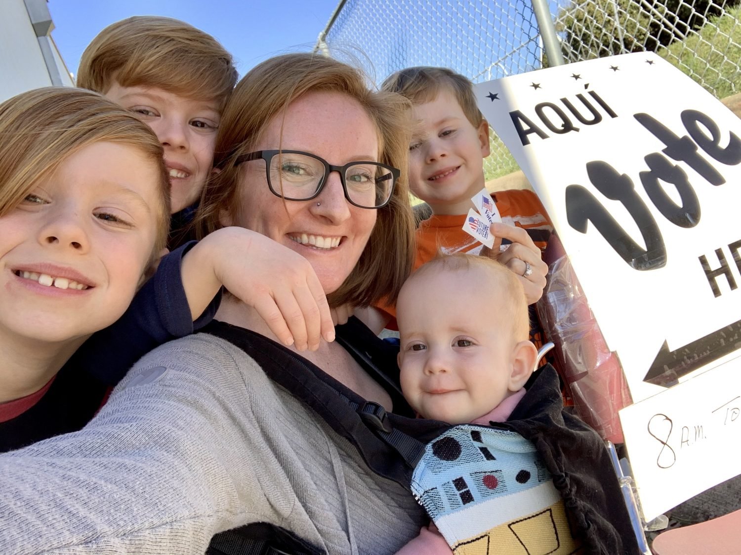 Parent wearing an infant in a carrier surrounded by 3 other preschool age children next to a sign saying “Aquí Vote Here” and they are holding “I voted” stickers.