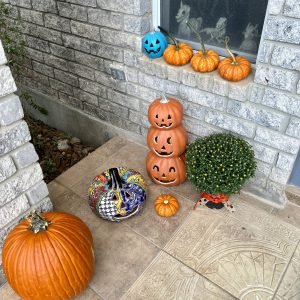Photo of front porch with mixture of real and artificial pumpkins. There is a teal pumpkin on the window seal next to orange pumpkins. 