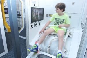 a boy sitting on a replica of a space toilet found in the space station
